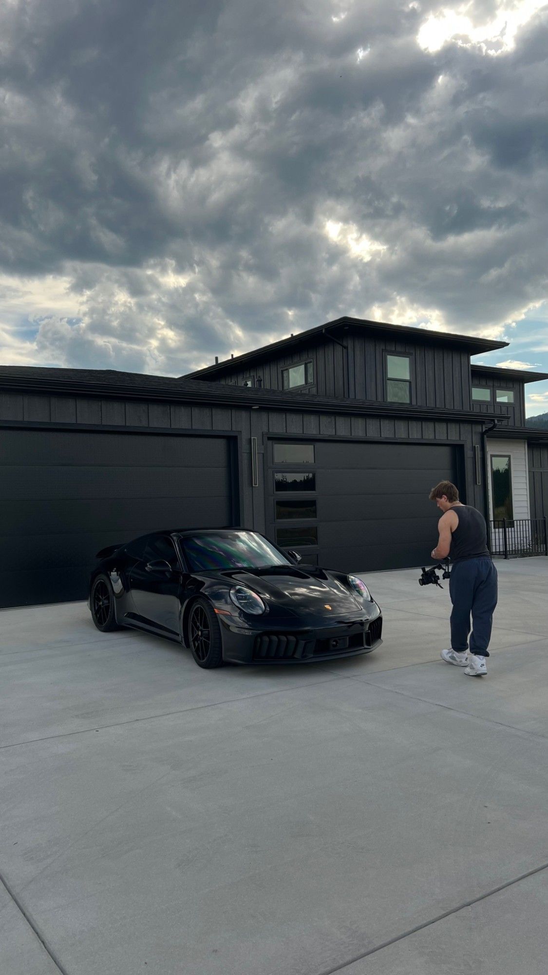 Gabe filming a black Porsche 911 with a gimbal camera in a driveway under storm clouds